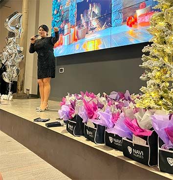 A woman in a sparkling black dress stands on a stage and speaks enthusiastically into a microphone at a company holiday party.