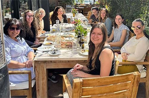A group of about ten professional women share a meal at a long wooden table on a sunny restaurant patio. The table is filled with dishes and drinks, and the women are smiling and enjoying a collegial lunch together.